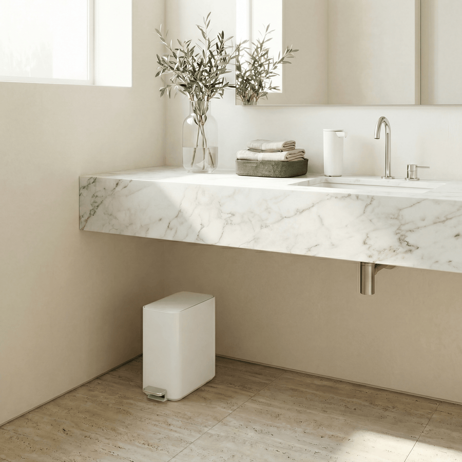 Simplehuman's 9L Slim Step Can in White in a very bright bathroom environment with white marble sink counter top and walls featuring a Simplehuman Sensor Pump in white on the countertop.